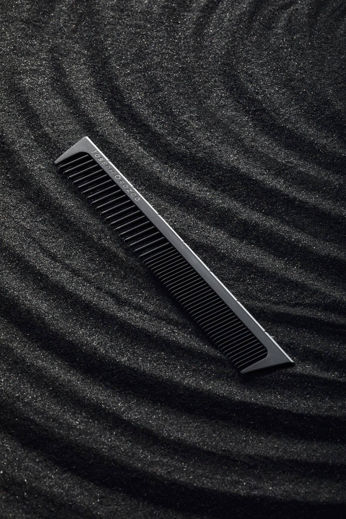 A professional hair comb from Joseph Orozco, designed for styling, rests diagonally on a textured, shimmering black surface that suggests the sleekness and elegance of the styling tools and products by this pro hair brand.