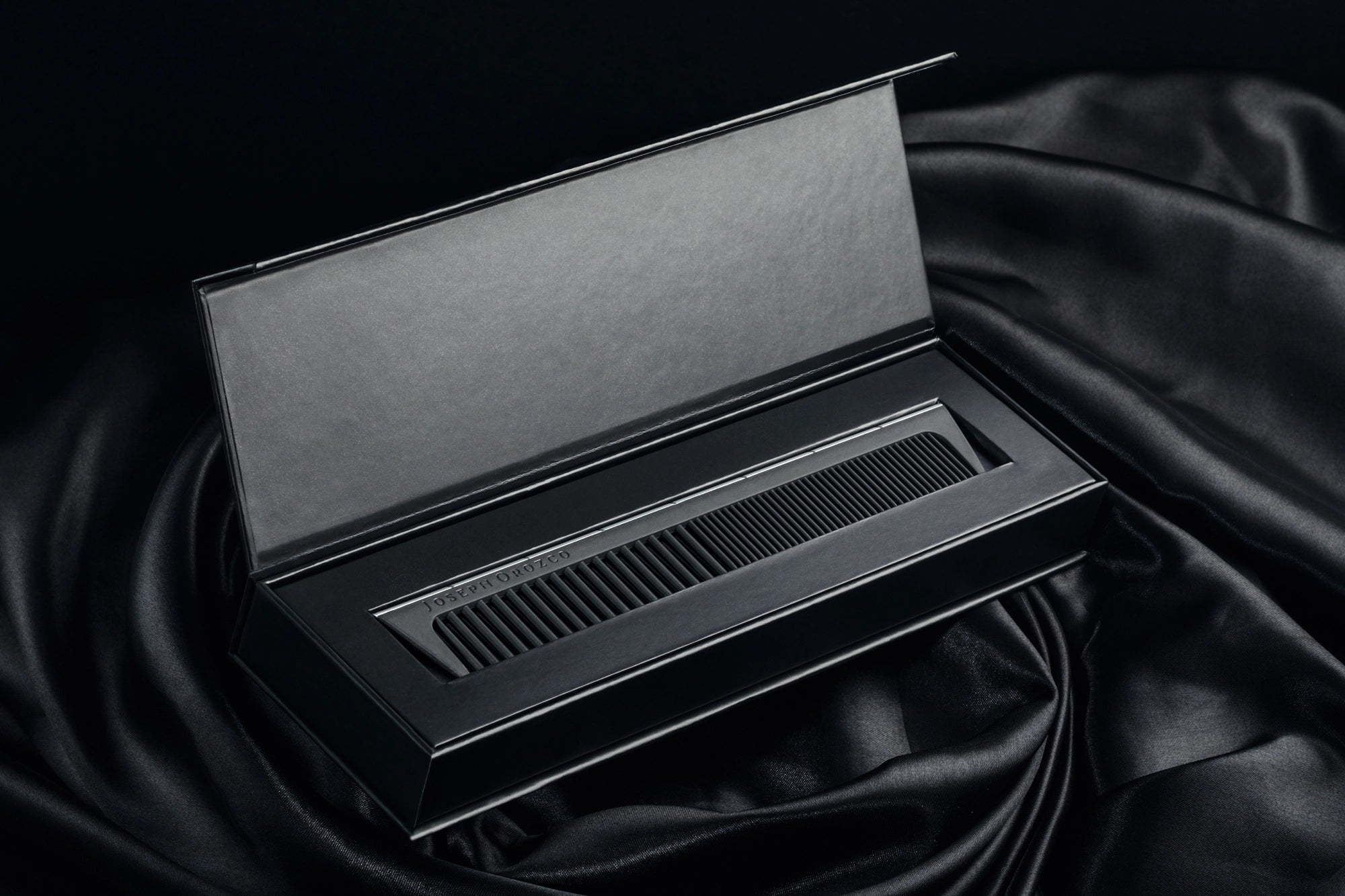 An elegant Joseph Orozco professional hair comb is presented within a sophisticated matte black box, placed on a luxuriously draped satin fabric, highlighting the premium quality of the hair styling tools by this pro hair brand.